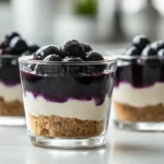 No-Bake Blueberry Cheesecake Cups with graham cracker crust, creamy cheesecake filling, and blueberry topping served in clear dessert cups in a bright modern American kitchen.