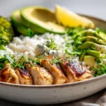 Healthy chicken rice bowl with avocado, broccoli, rice, cucumber, and lemon yogurt sauce on a white kitchen counter.